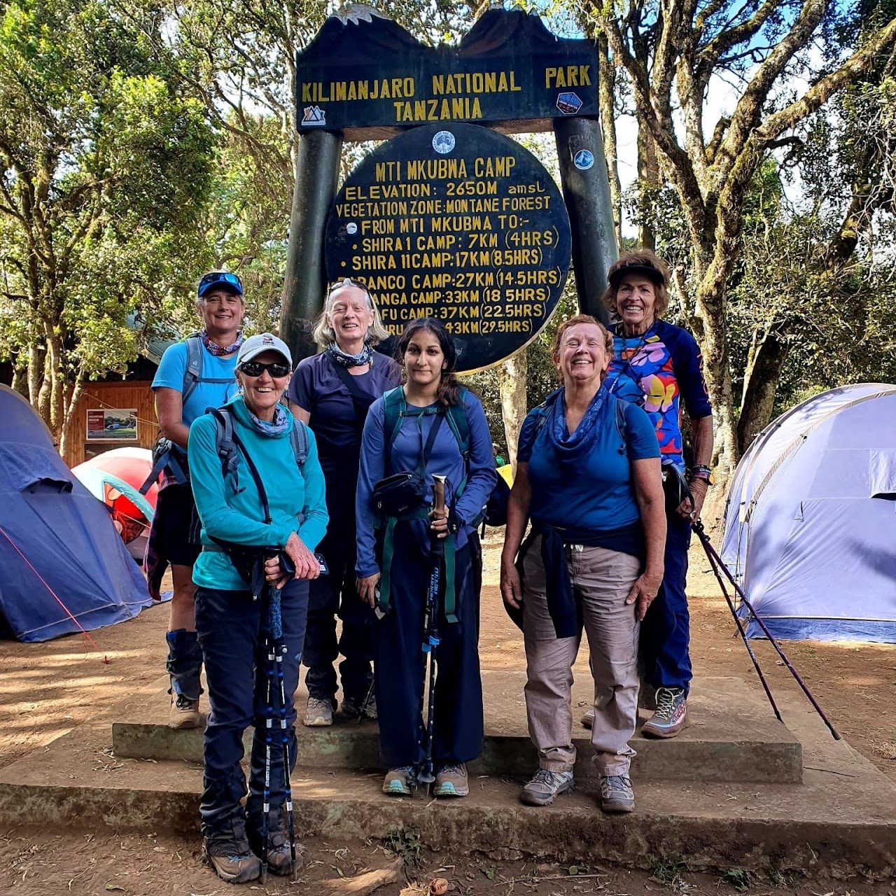 Six smiling women at the Mti Mkubwa Camp sign in the montane forest