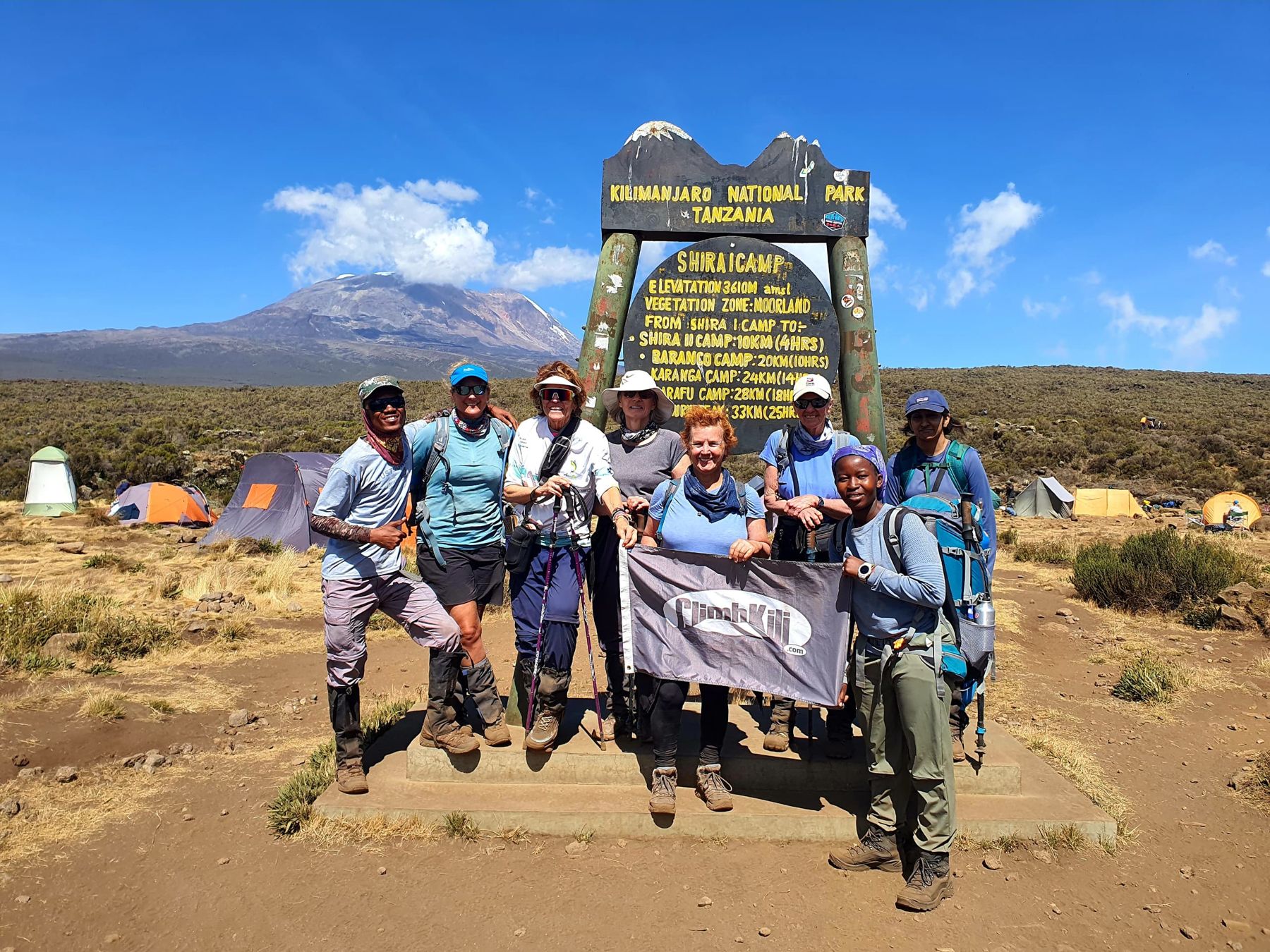 The group holding the Climb Kili flag at the Shira Camp sign with Kibo in the distance