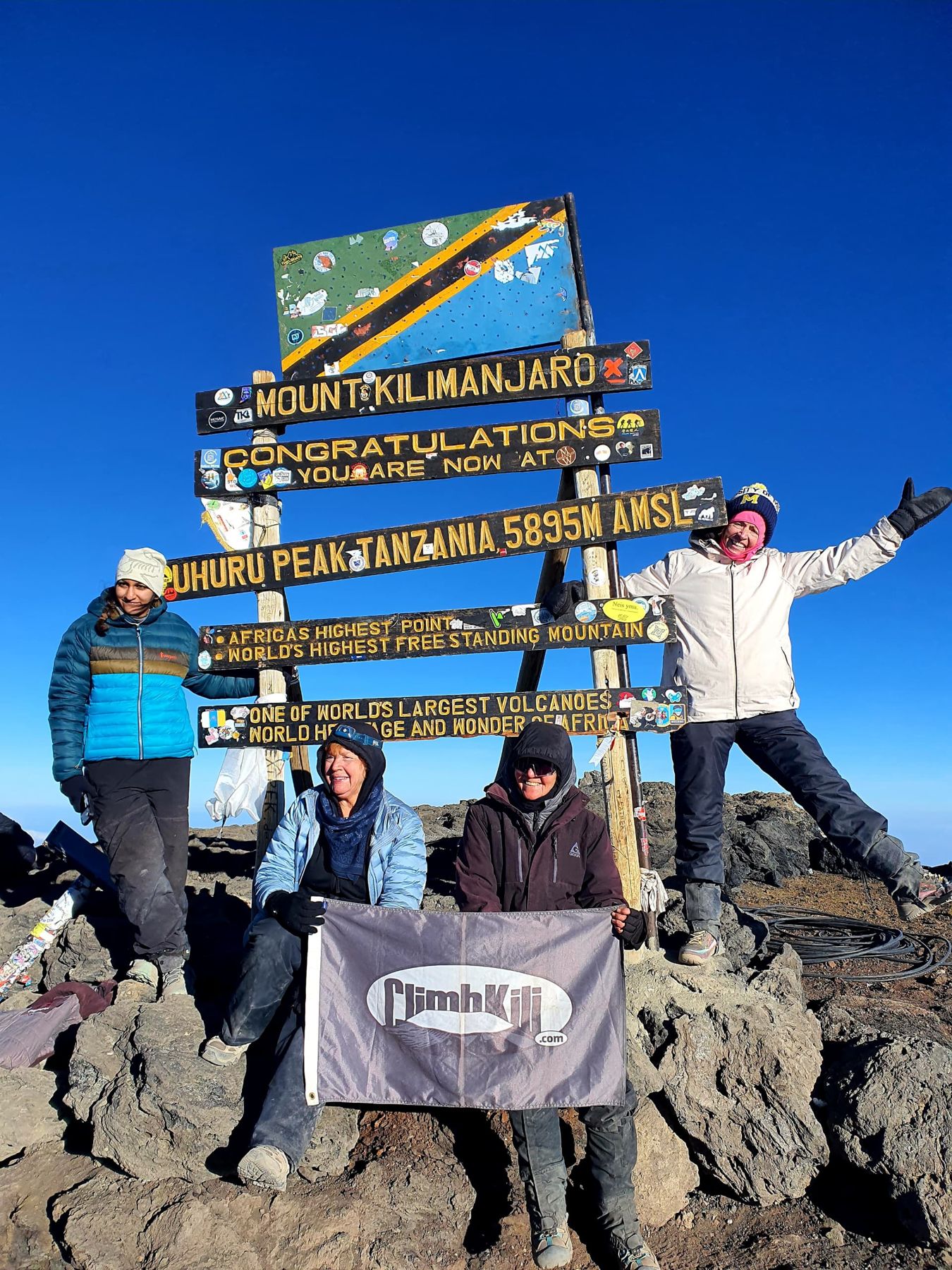 Four women celebrating with the Climb Kili flag at the Uhuru Peak summit sign